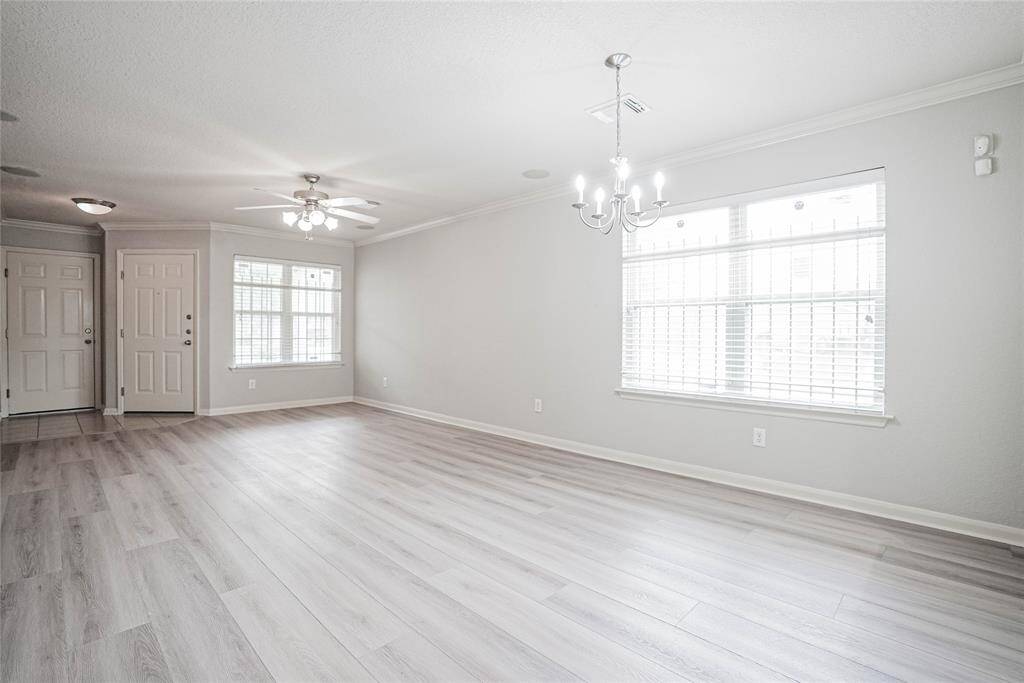 Bright and spacious living area with light wood flooring, large windows for natural light, and modern lighting fixtures. Fresh and inviting atmosphere with neutral walls and a ceiling fan for comfort.