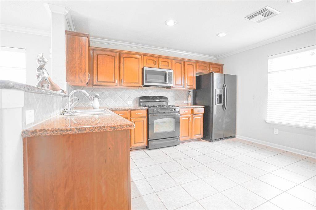 Bright kitchen with tiled floors, granite countertops, and wooden cabinetry. Features stainless steel appliances, including a gas stove and double-door refrigerator. Ample natural light from a large window.