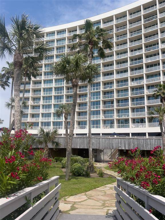 This photo showcases a modern high-rise building with numerous balconies, surrounded by lush landscaping and palm trees. A stone path and wooden bridge lead up to the entrance, offering a welcoming, tropical vibe.