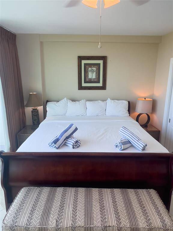 This inviting bedroom features a large bed with elegant wooden frame, complemented by tasteful lamps on bedside tables. Natural light filters through the curtains, creating a cozy and serene atmosphere. A patterned bench at the foot of the bed adds extra charm.