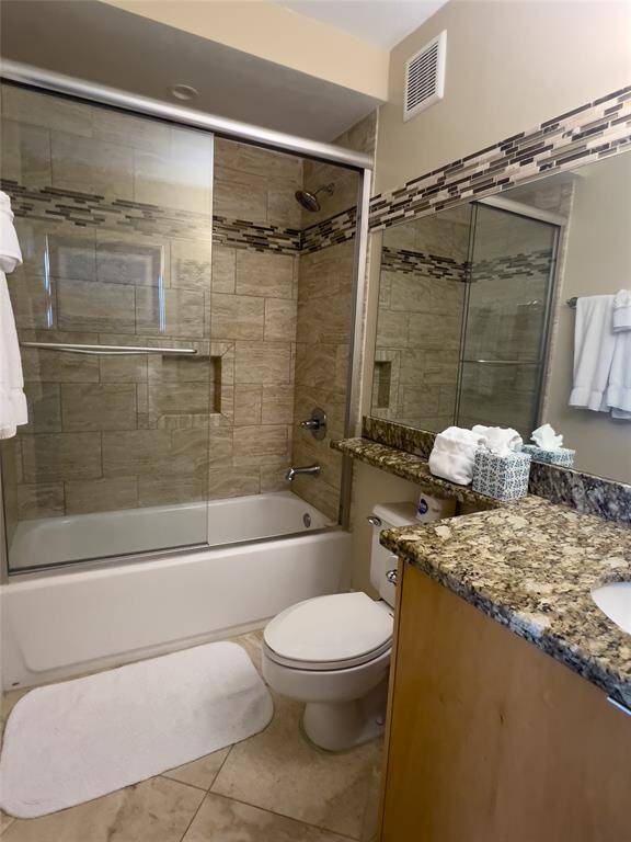 This bathroom features a modern design with a tiled shower and tub combo, glass doors, and a granite countertop. It also includes a large mirror, ample lighting, and neutral tones for a warm, inviting feel.