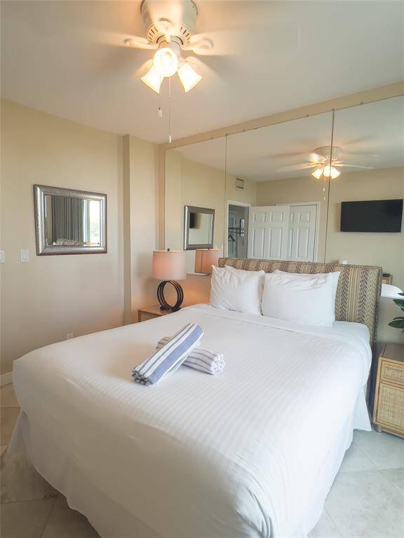 This cozy bedroom features a comfortable bed, mirrored wall for added space perception, ceiling fan, and natural light. It includes a corner TV and a stylish bedside lamp, offering a relaxing and practical space.