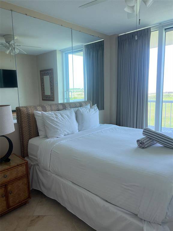 This cozy bedroom features a comfortable bed, mirrored wall, and large windows with drapes offering plenty of natural light. The room includes a ceiling fan, wall-mounted TV, and a bedside table with a lamp, creating a relaxing and inviting atmosphere.