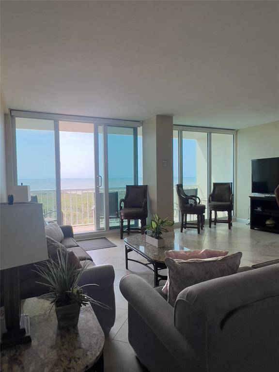 Bright living room with large windows offering ocean views. Features include tiled floors, comfortable seating, and a TV. Ideal for relaxation and entertaining.