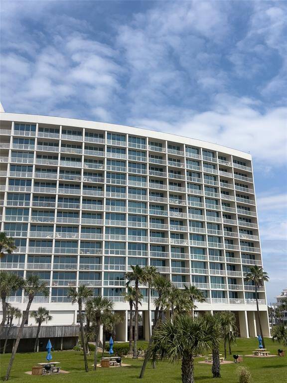 Modern high-rise condominium with expansive glass balconies, surrounded by well-maintained green spaces and palm trees, under a clear blue sky.
