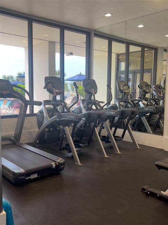 This photo showcases the building gym with large windows, featuring cardio equipment like treadmills and elliptical machines, and offering views of an outdoor pool area.