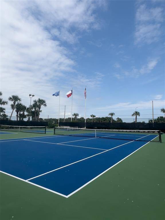 Tennis anyone?  This photo features a well-maintained tennis court with a blue surface, surrounded by palm trees and three flags. The setting suggests a coastal or resort-like environment, perfect for recreational activities.