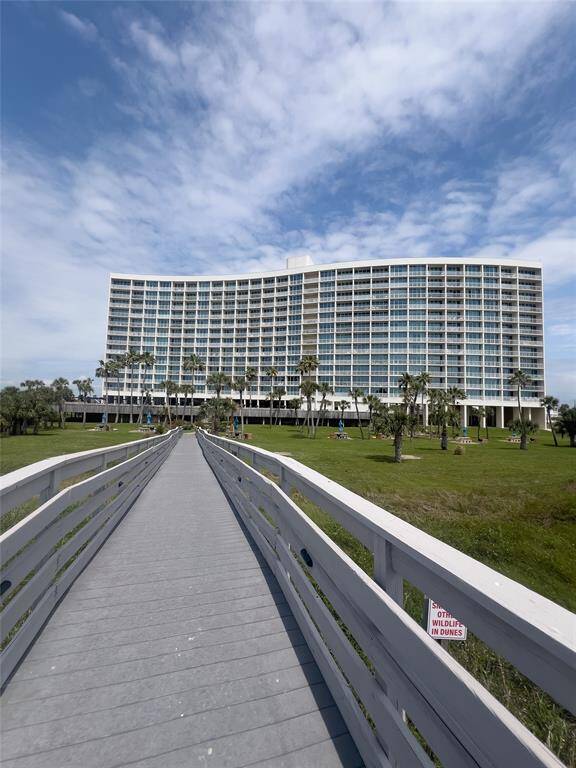 This photo showcases a large, modern beachfront condominium with extensive glass windows and a sleek design. The building is surrounded by a well-maintained grassy area and is accessible via a long boardwalk, offering scenic views and a coastal lifestyle.