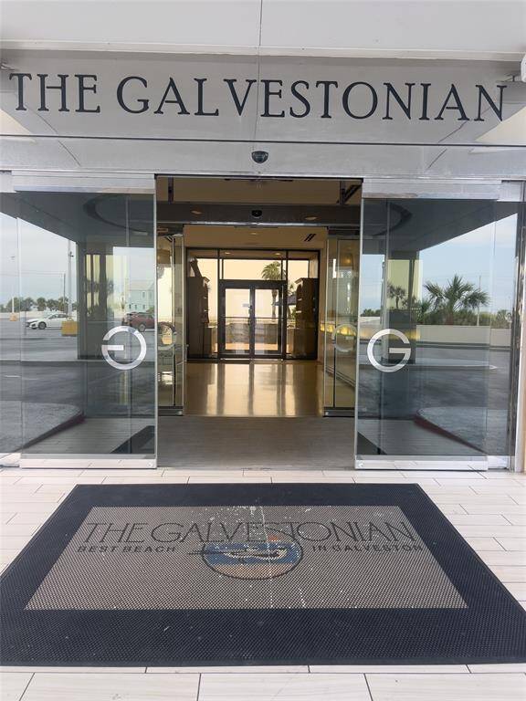 Modern entrance to "The Galvestonian," featuring glass doors and a welcoming mat. Ideal for those seeking a stylish beachfront living experience.