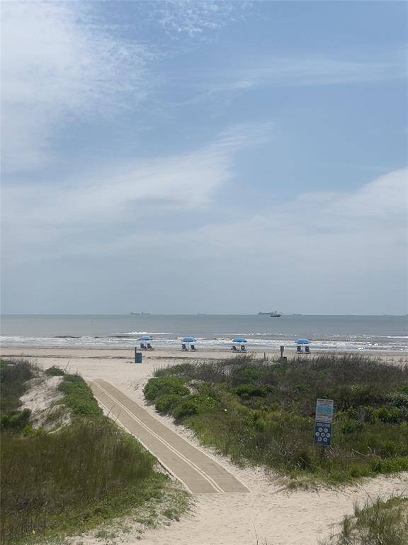 Tranquil beach view with a sandy path leading to the ocean, lined with blue umbrellas and lush greenery, perfect for seaside relaxation.