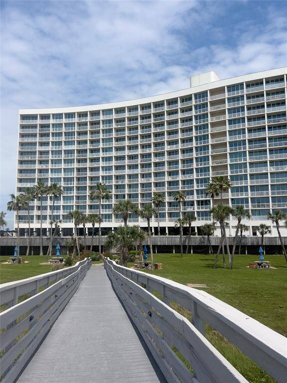 Modern high-rise building with extensive glass windows, set against a clear sky. A boardwalk leads to the entrance, surrounded by well-maintained green lawns and palm trees, creating a coastal and inviting atmosphere.