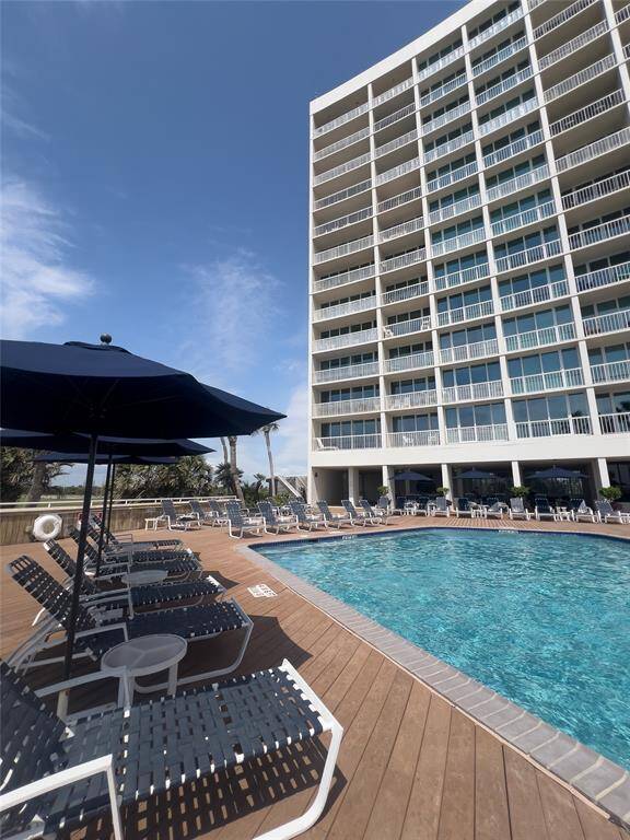 This photo showcases a modern high-rise building with spacious balconies and a large, inviting outdoor pool area. The pool is surrounded by a wooden deck with sun loungers and umbrellas, perfect for relaxation. The setting is bright and open, ideal for enjoying sunny days.