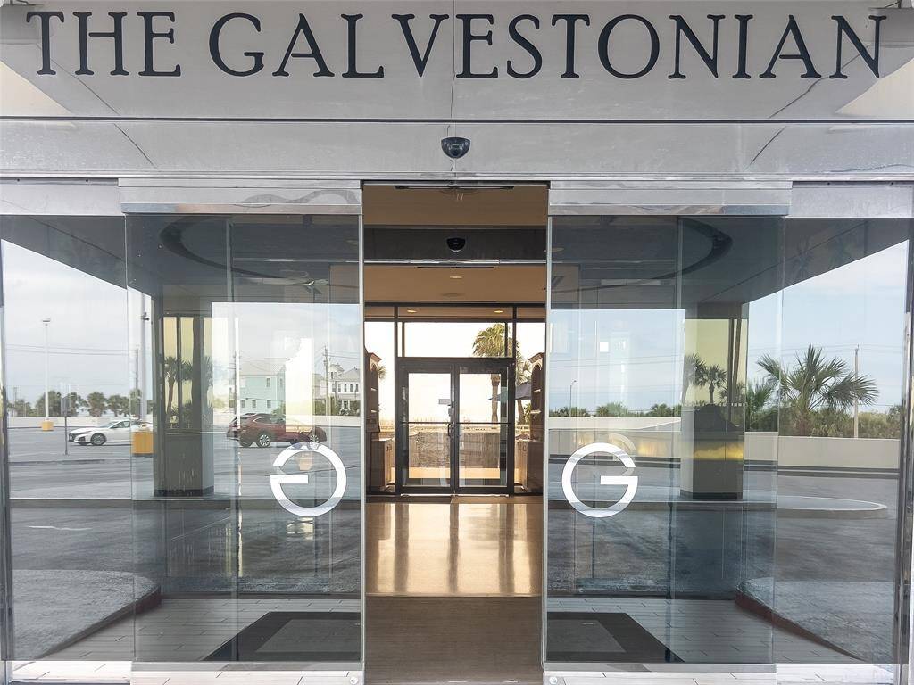 Modern entrance to The Galvestonian, featuring sleek glass doors with a view of the surroundings, including palm trees and nearby buildings.