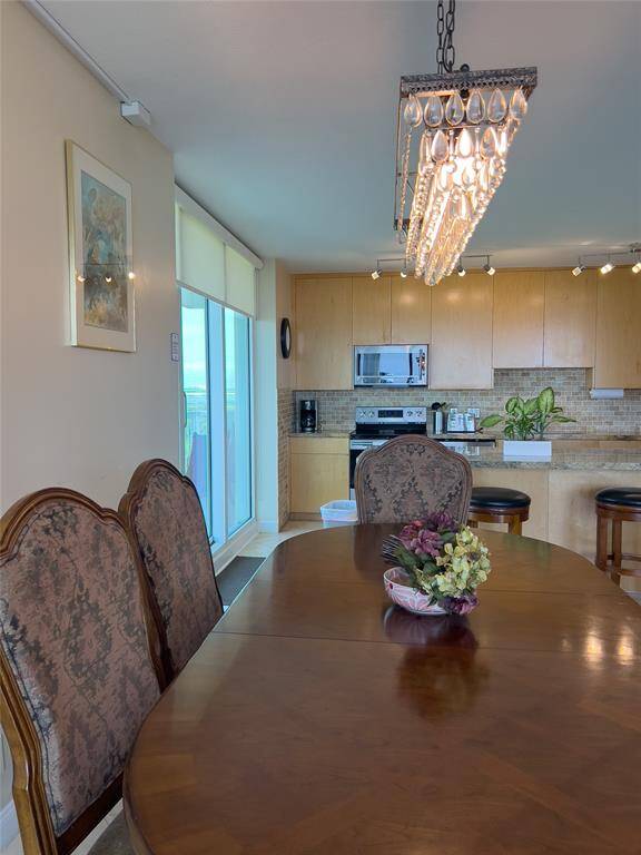 This dining area features an elegant chandelier above a wooden table with upholstered chairs. It leads into a modern kitchen with light wood cabinets, stainless steel appliances, and a breakfast bar. Large windows offer ample natural light.