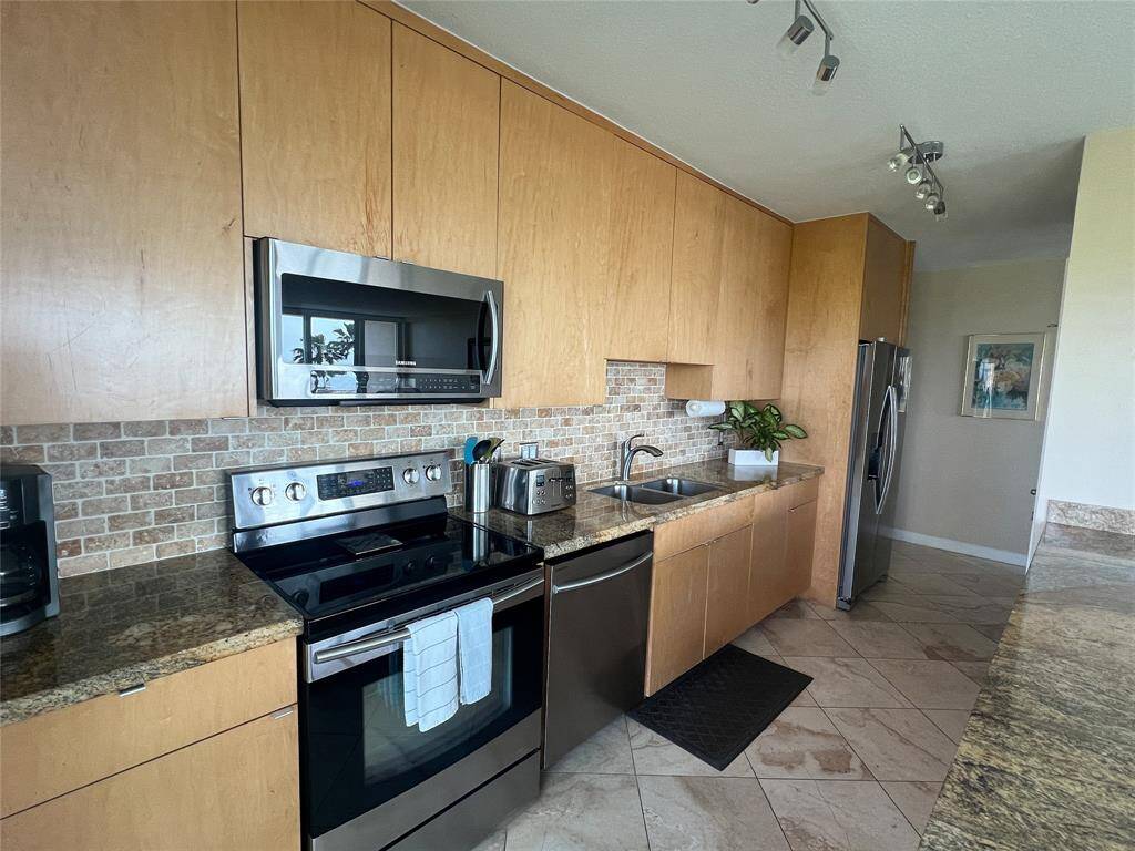 Modern kitchen with light wood cabinetry, granite countertops, and stainless steel appliances. Features include a sleek tile backsplash, double sink, and ample storage. Bright and inviting space with tiled flooring and track lighting.
