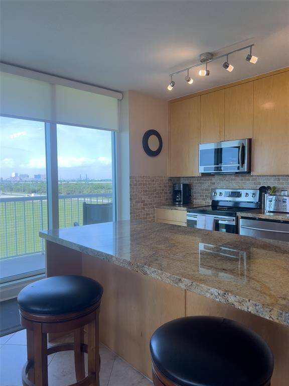 Modern kitchen with stainless steel appliances, light wood cabinetry, and granite countertops. Features a large island with seating and track lighting. Offers stunning views through expansive windows.