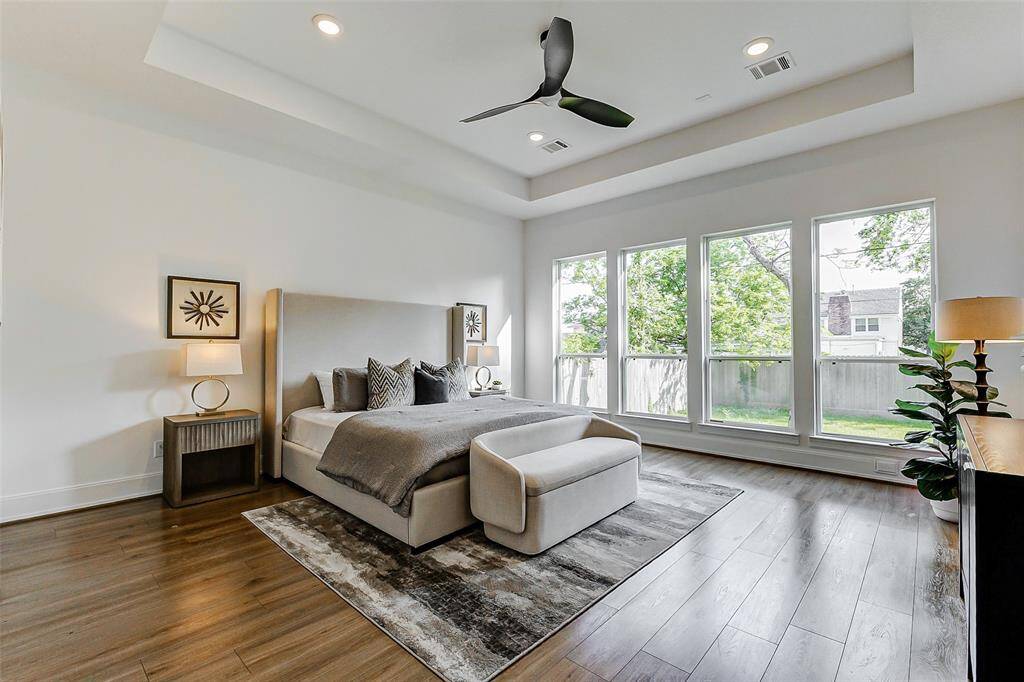 Retreat to the serene comfort of the Owner’s Suite, featuring a dramatic tray ceiling, a sleek ceiling fan, recessed lighting, and a luxurious en suite bathroom. A wall of picture windows bathes the room in natural light, creating a peaceful sanctuary.