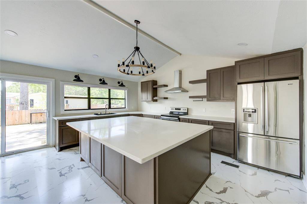 This modern kitchen features a spacious open layout with a large island, stainless steel appliances, and sleek cabinetry. Natural light streams through large windows, and the sliding door provides patio access. Elegant marble-style flooring and a contemporary chandelier add to its charm.