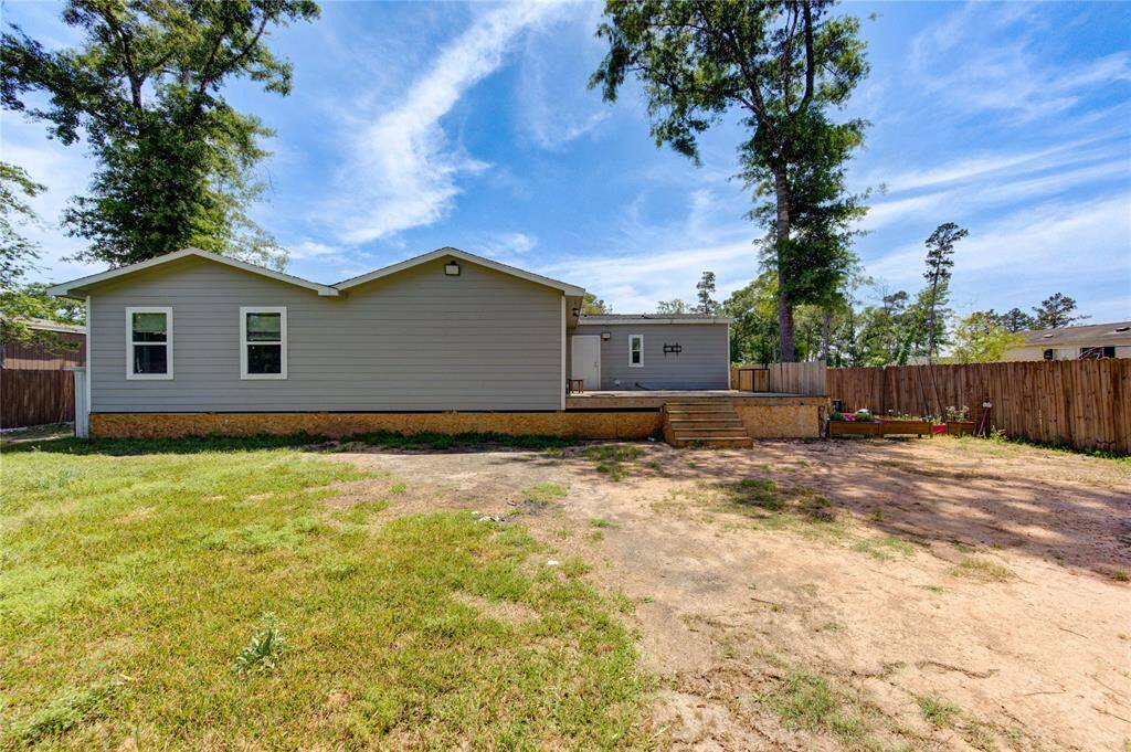 This photo shows the backyard of a single-story home with a spacious deck, surrounded by a wooden fence. There's a mix of grass and bare soil, with a few mature trees providing shade. The area offers potential for landscaping or outdoor activities.
