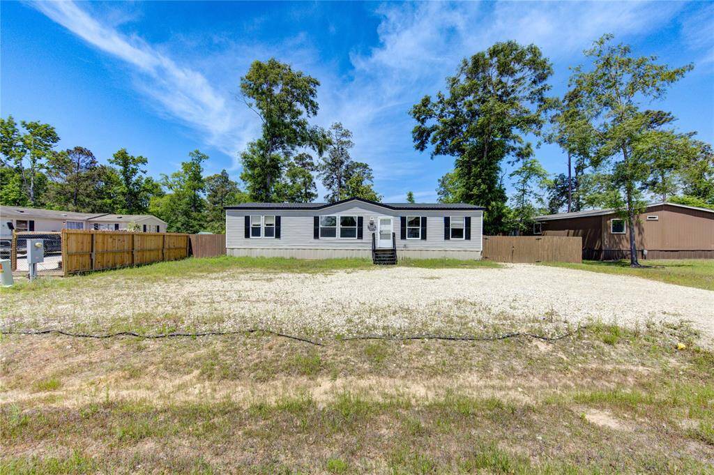 This property features a spacious manufactured home set back from the street, surrounded by a large yard with mature trees. The exterior is well-maintained, featuring a neutral color palette. A gravel driveway provides ample parking space, and the area is fenced for added privacy.