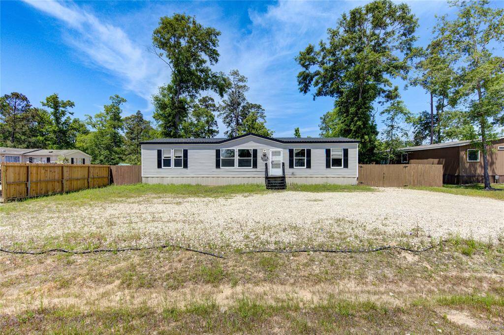 This photo shows a single-story, modular home with a light exterior and dark shutters, situated on a spacious lot with a gravel driveway. The yard is partially fenced, and the property is surrounded by mature trees, providing a sense of privacy.