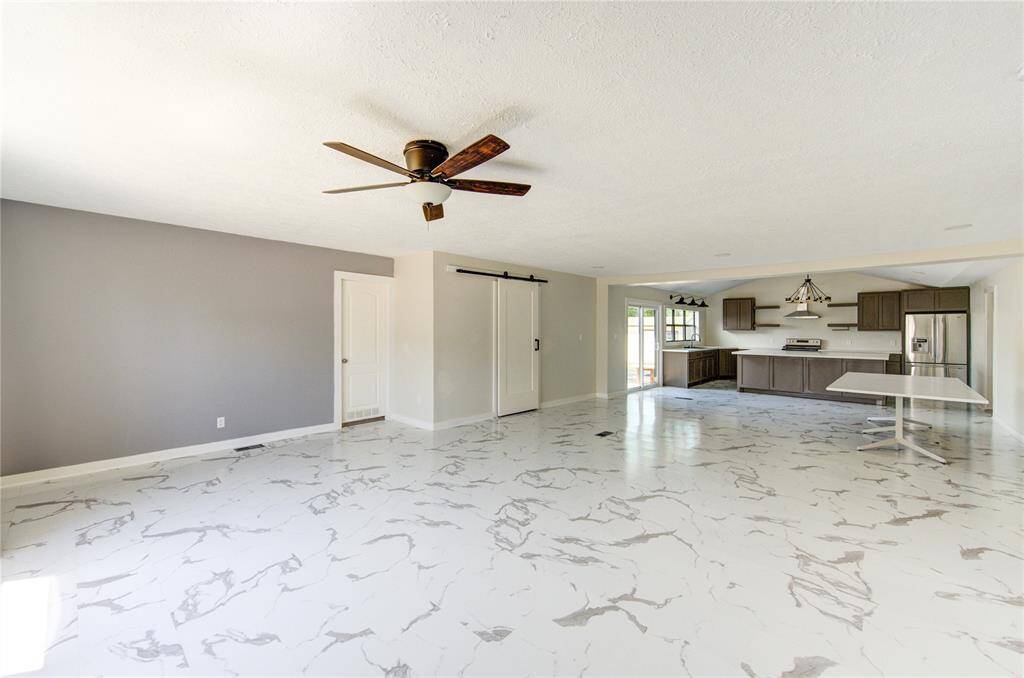 Spacious open-concept living area with modern kitchen. Features include marble-like flooring, a ceiling fan, a sliding barn door, and ample natural light. Ideal for entertaining and family gatherings.
