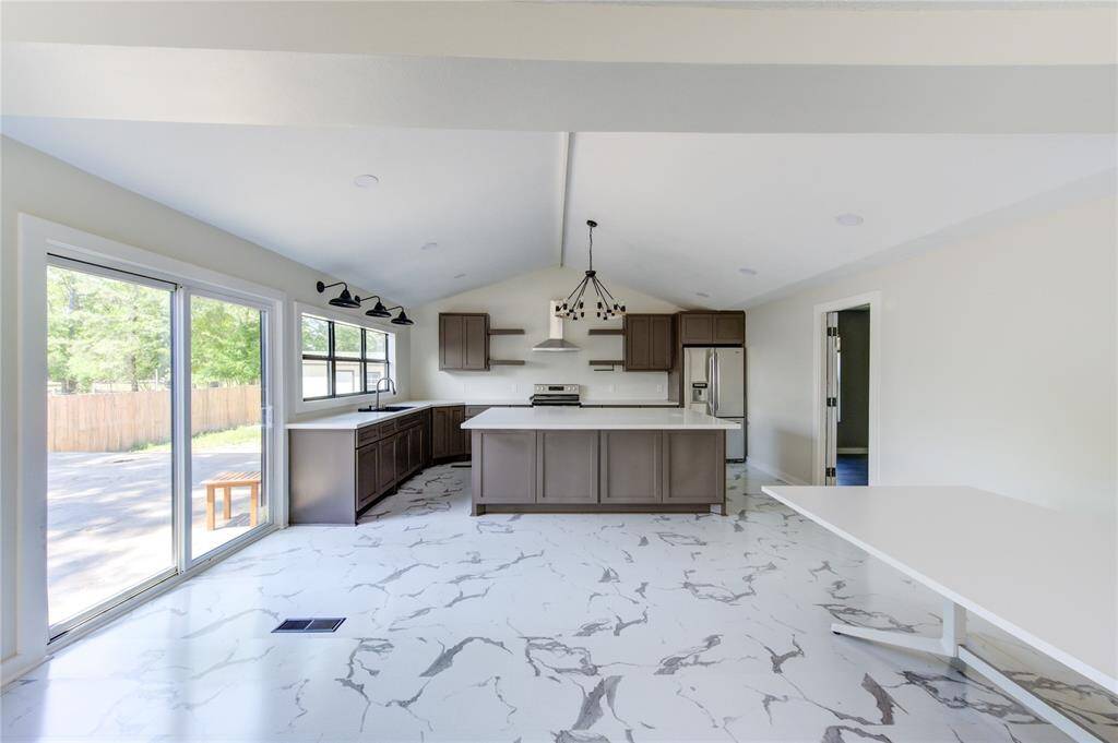 This spacious kitchen features modern cabinetry, marble-patterned flooring, and a large island. It has abundant natural light from sliding glass doors and windows, with stylish pendant lighting and stainless steel appliances. Ideal for cooking and entertaining.