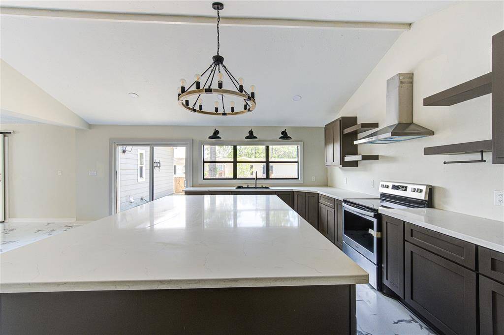 This kitchen features a spacious layout with a large island, modern dark cabinetry, and stainless steel appliances. The room is well-lit with natural light from large windows and a sliding door, complemented by a stylish chandelier and pendant lights.