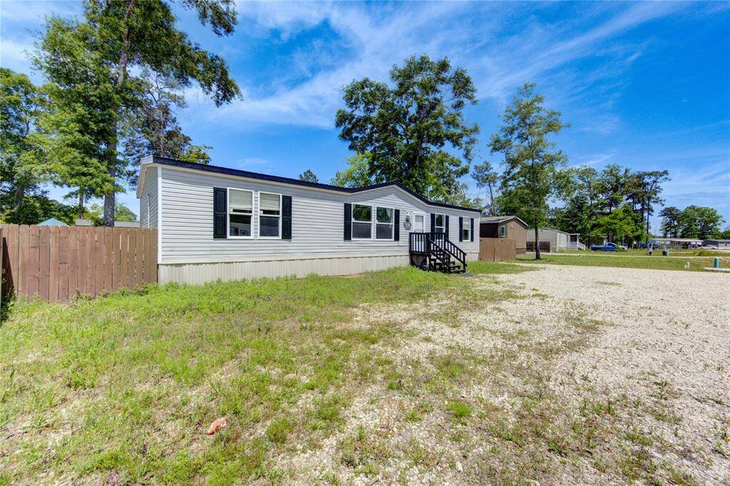 This photo shows a single-story manufactured home with a white exterior and black shutters, set on a spacious lot. It features a small front porch, a gravel driveway, and a fenced yard, surrounded by trees for added privacy.