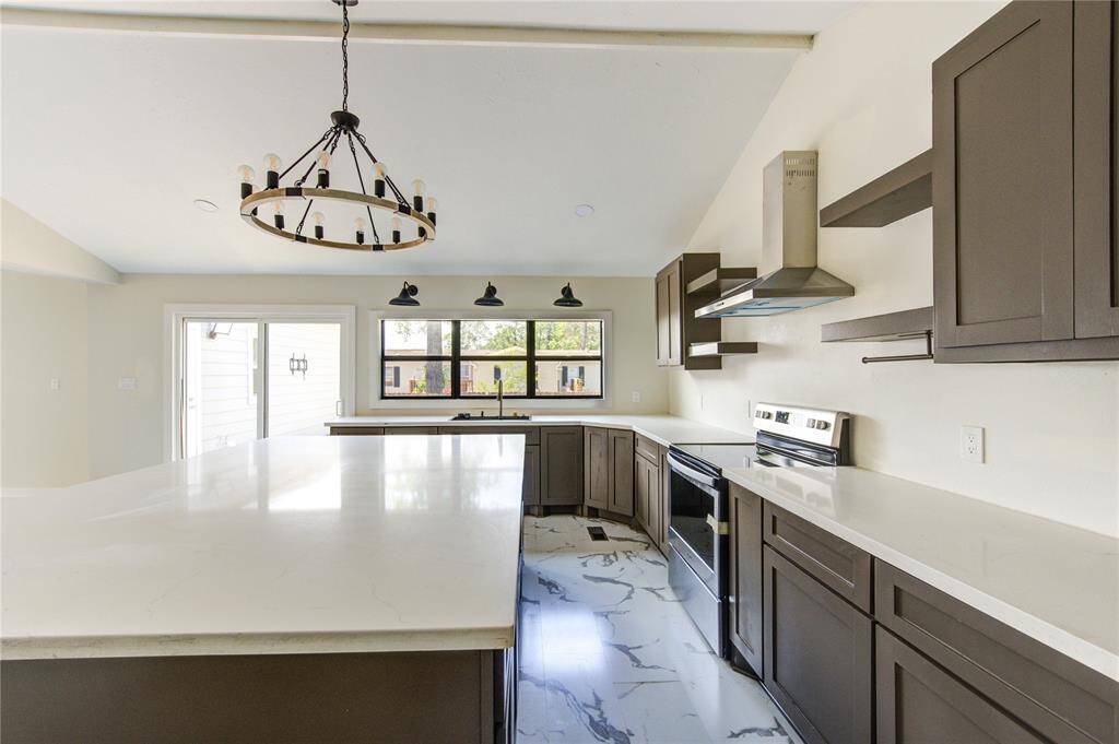 This modern kitchen features sleek dark cabinetry, a spacious marble island, and stainless steel appliances. Large windows and a sliding glass door offer ample natural light, while a stylish chandelier adds elegance. Perfect for cooking and entertaining.