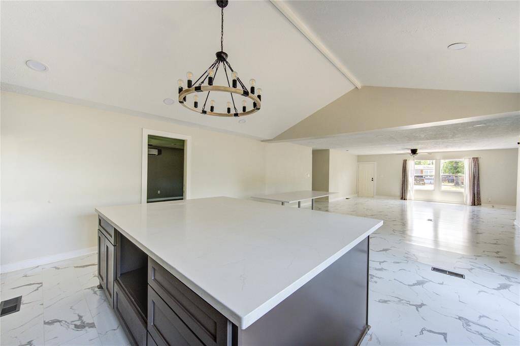 This spacious open-concept area features a modern kitchen with a large island, sleek countertops, and ample cabinet space. The room is bright with natural light from large windows, complemented by elegant tile flooring and a stylish chandelier. Perfect for entertaining or family gatherings.