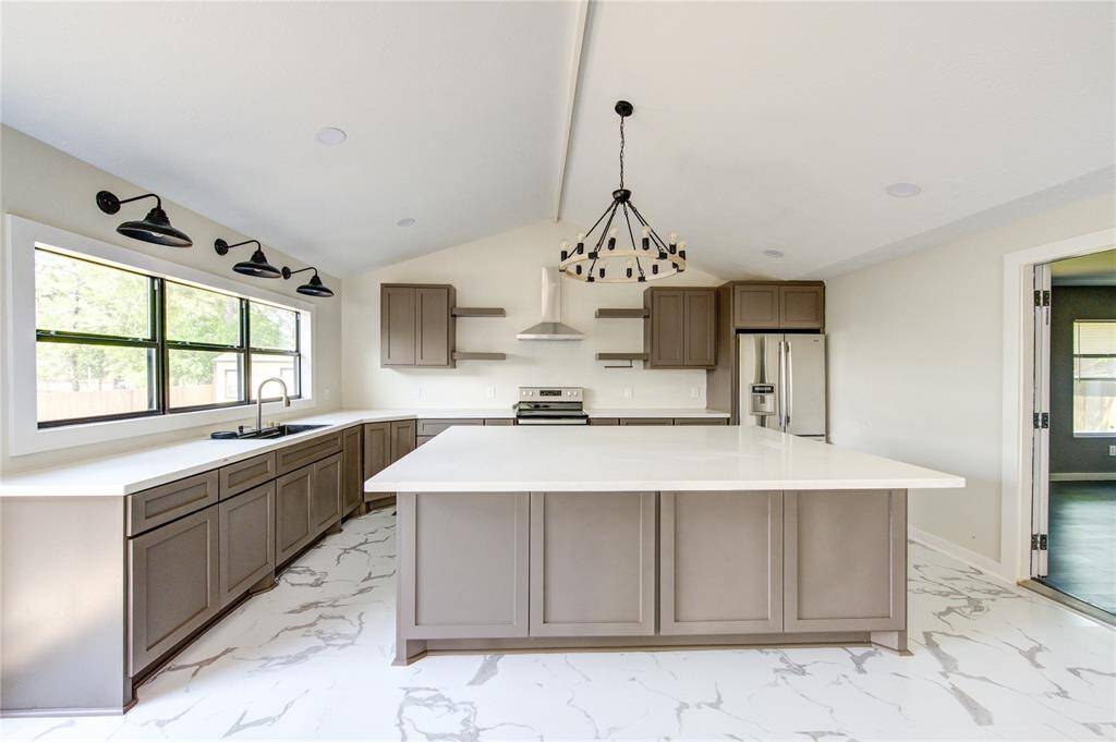 This modern kitchen features sleek gray cabinets, a large central island with white countertops, and stainless steel appliances. It has a vaulted ceiling, stylish lighting fixtures, and ample natural light from large windows, complemented by marble-patterned flooring.