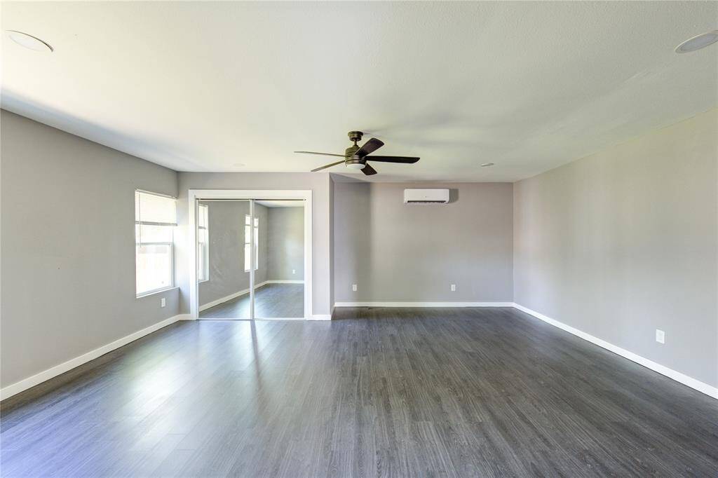 This spacious room features sleek dark wood flooring, neutral gray walls, and ample natural light from two windows. It includes a ceiling fan, mirrored closet doors, and a wall-mounted air conditioning unit for comfort and convenience.