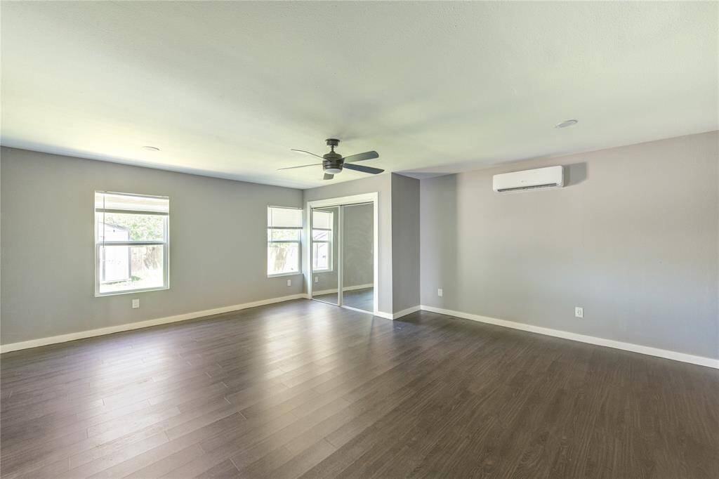 Spacious living room with hardwood floors, neutral walls, and ample natural light from multiple windows. Features a ceiling fan, wall-mounted air conditioning unit, and sliding glass doors.