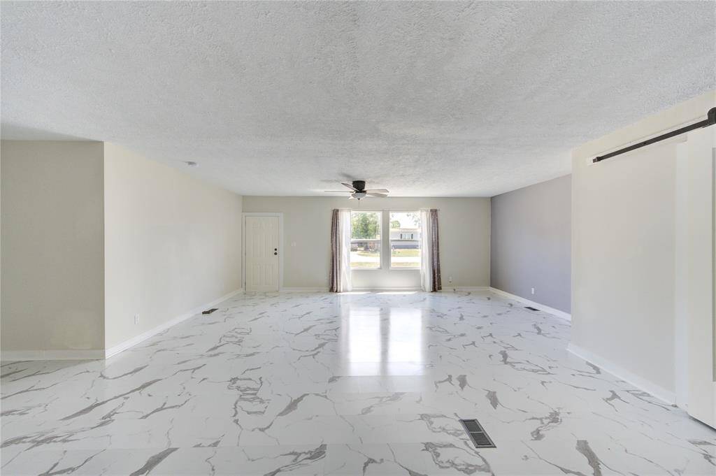 This spacious living area features elegant marble-patterned tile flooring, neutral walls, and ample natural light from large windows. A ceiling fan provides comfort, and the room offers versatile layout options.