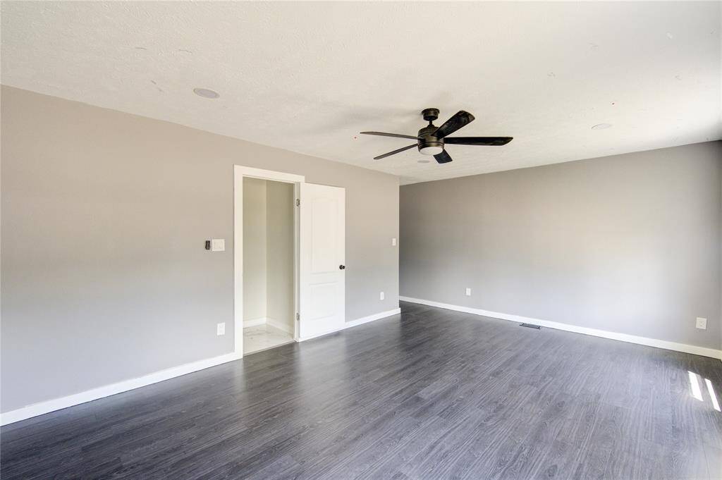 This room features a spacious, light-filled area with modern gray walls and dark wood flooring. It includes a ceiling fan and a door leading to another space, offering a clean and versatile backdrop for personal touches.