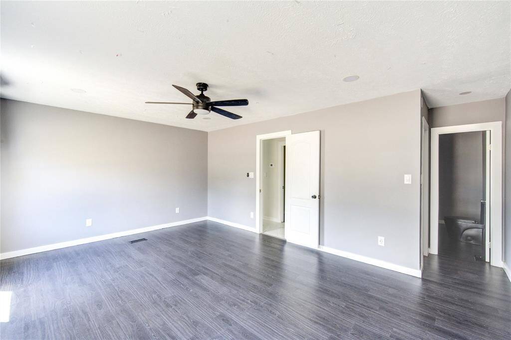 This room features sleek dark wood flooring, light gray walls, and a ceiling fan. It has a modern, open feel with direct access to a bathroom and another adjacent space.