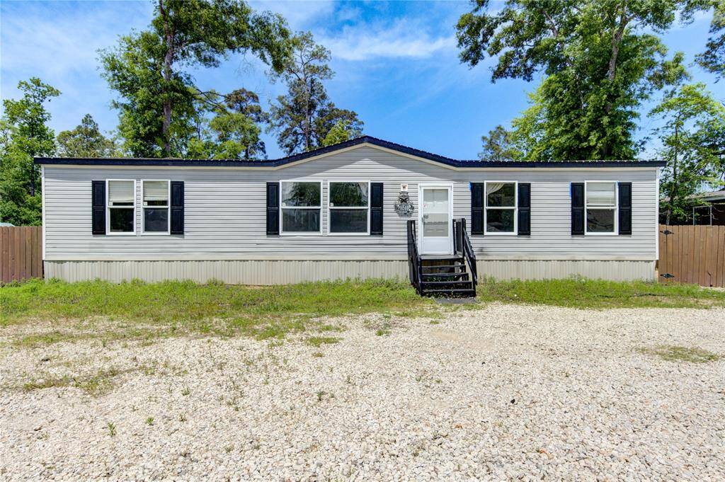 This is a single-story manufactured home with a light gray exterior, black shutters, and a central front entrance. It features a gravel driveway and is surrounded by trees, providing a private setting.