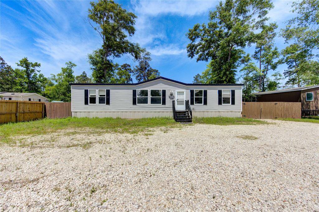This photo features a single-story home with a white exterior and black shutters. It has a small front porch with steps leading to the entrance. The yard is spacious with a gravel driveway and is bordered by wooden fencing, surrounded by mature trees for added privacy.