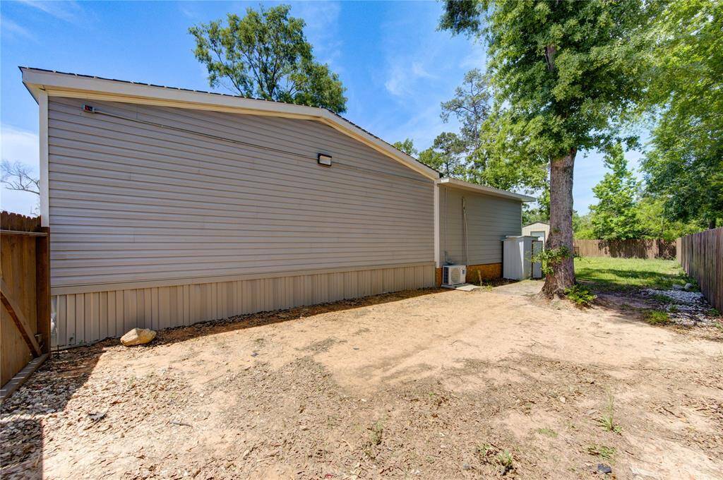 This photo shows the side yard of a home with a beige exterior, bordered by a wooden fence. The area is mostly unpaved with some grass and trees, offering a private outdoor space.