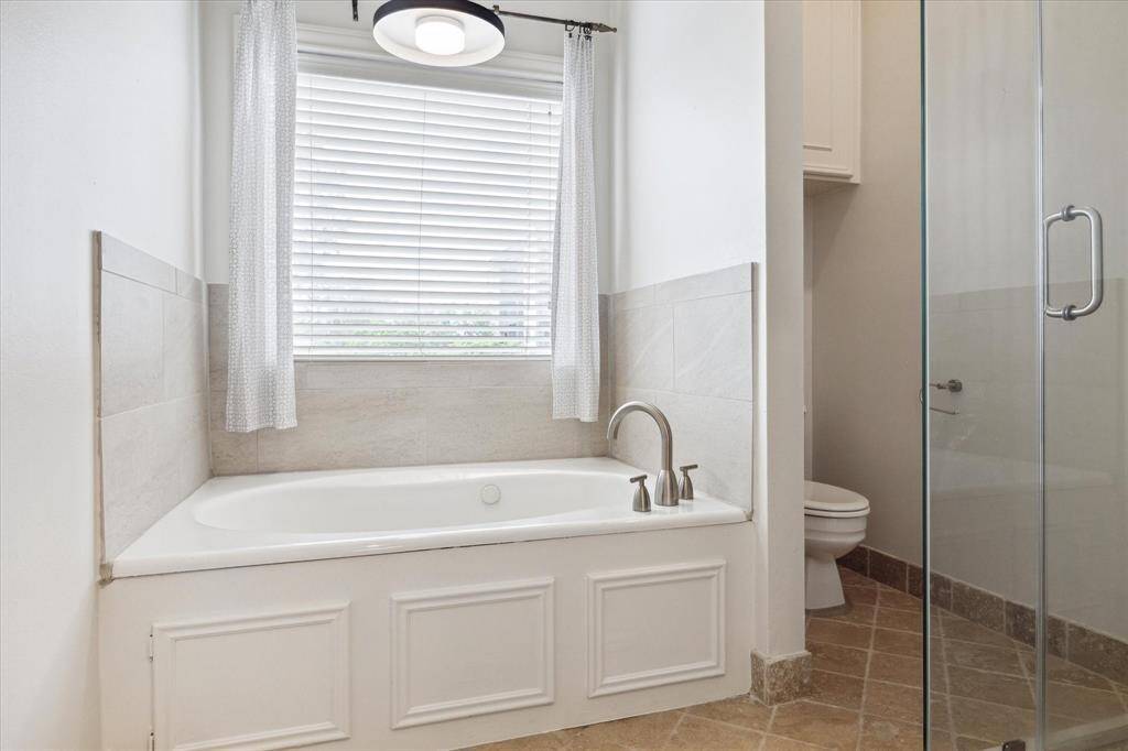 The Primary Bath features a clean, modern design with a built-in jetted bathtub under a window, a separate glass-enclosed shower, and neutral tile flooring.