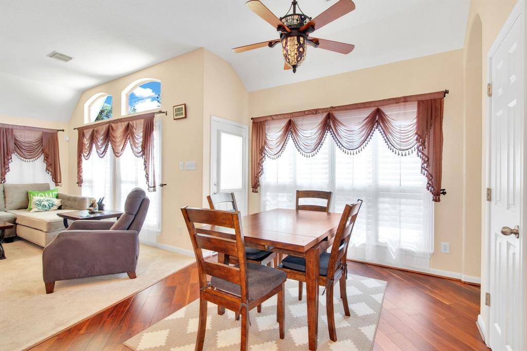 This breakfast area features large windows with elegant drapery, allowing plenty of natural light, with warm wood flooring in the dining section and a ceiling fan.