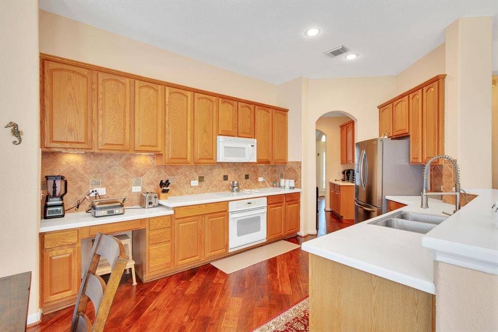 This kitchen features warm wooden cabinetry, white countertops, modern appliances, and tile backsplash. The open layout includes ample storage and a double sink, with hardwood floors adding elegance.