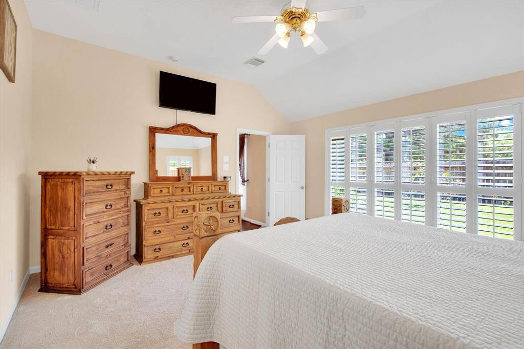 Bright and spacious primary bedroom with large windows featuring plantation shutters, providing plenty of natural light. It includes a ceiling fan, wooden furniture, and neutral walls, creating a cozy and inviting atmosphere.