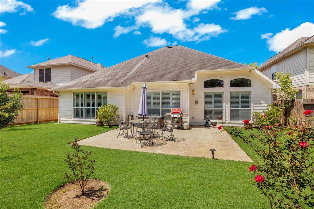 Charming backyard with a spacious patio area, perfect for outdoor dining and entertaining. The well-maintained lawn is bordered by wooden fencing, and the house features large windows.