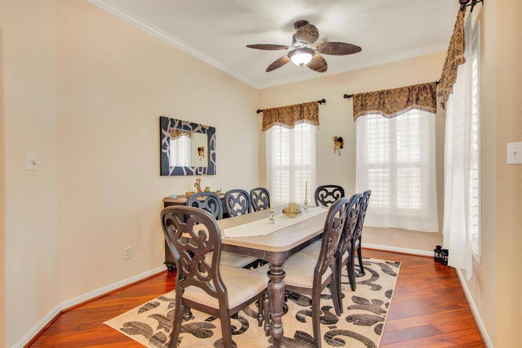 This is a bright, inviting dining room with warm wood flooring, a stylish ceiling fan, and large windows featuring elegant drapery.