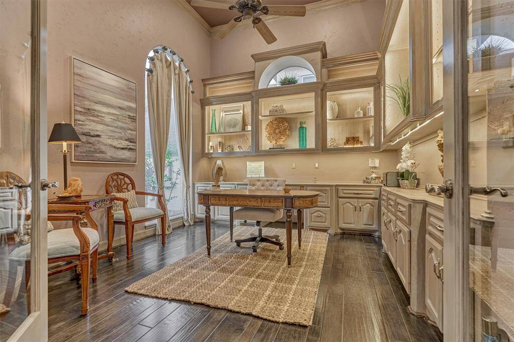 The spacious private office features a high ceiling with crown molding, custom built-ins with accent lighting, an arched window that floods the space with natural light, and rich wood flooring that adds warmth and sophistication.