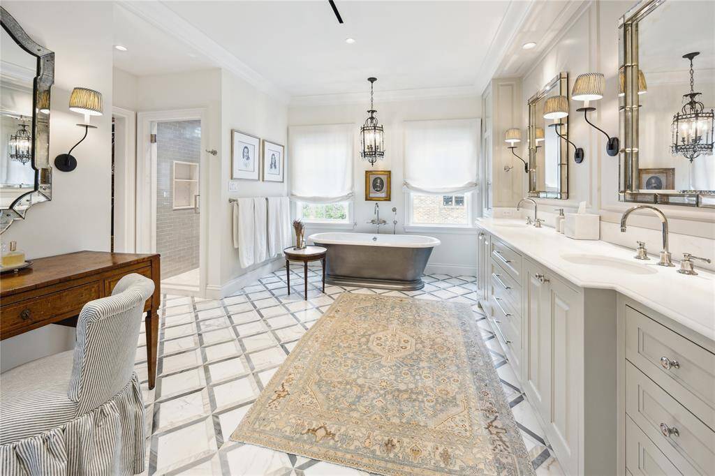 This luxurious Primary Bathroom showcases a freestanding soaking tub with a 18th century Italian chandelier above, Roman shades, and 3D geometric mosaic balmoral tile from Ann Sacks. There is also a vanity fashioned from an antique Italian Louis XVI style walnut desk accented by an antique Venetian mirror hanging above.