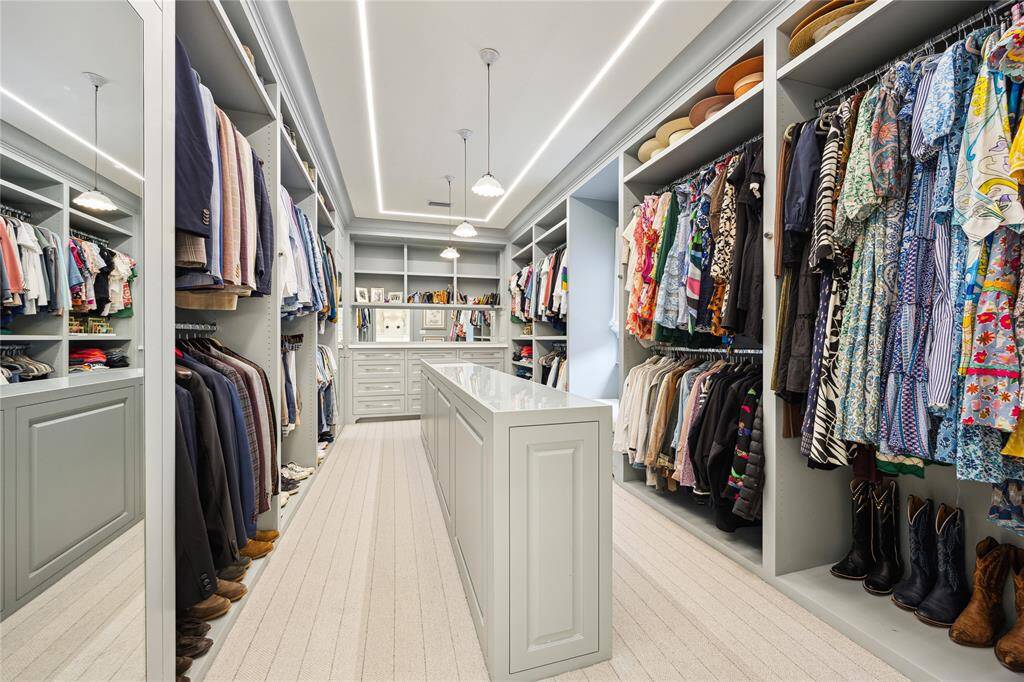 This expansive walk-in Closet features custom millwork and built-ins, ample hanging space, and a sleek island with integrated shoe storage, all illuminated by elegant pendant lighting and modern recessed accents.