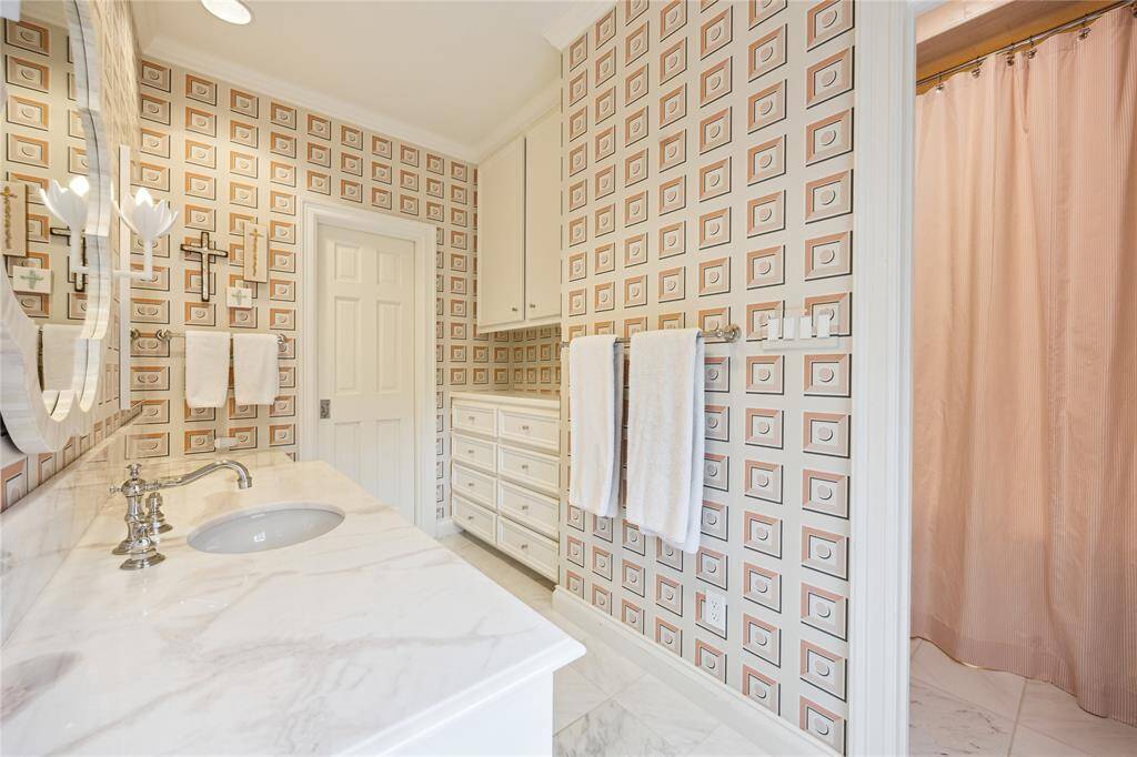 This stylish Bathroom features Schumacher Marcel Blush wallpaper, elegant marble countertops, ample built-in storage, and a walk in closet.